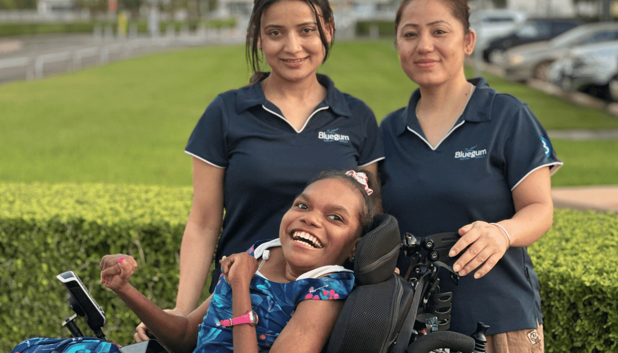 Stella smiling in her power wheelchair with two Bluegum support workers in Darwin.
