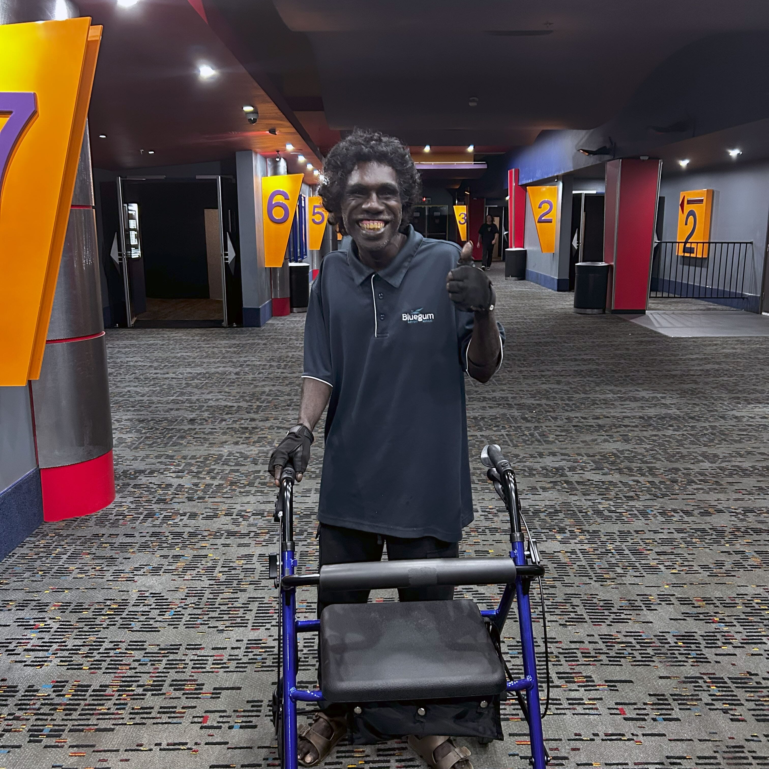 Jasco in a cinema foyer in Darwin with his walker, smiling and giving a thumbs up during a respite movie outing.