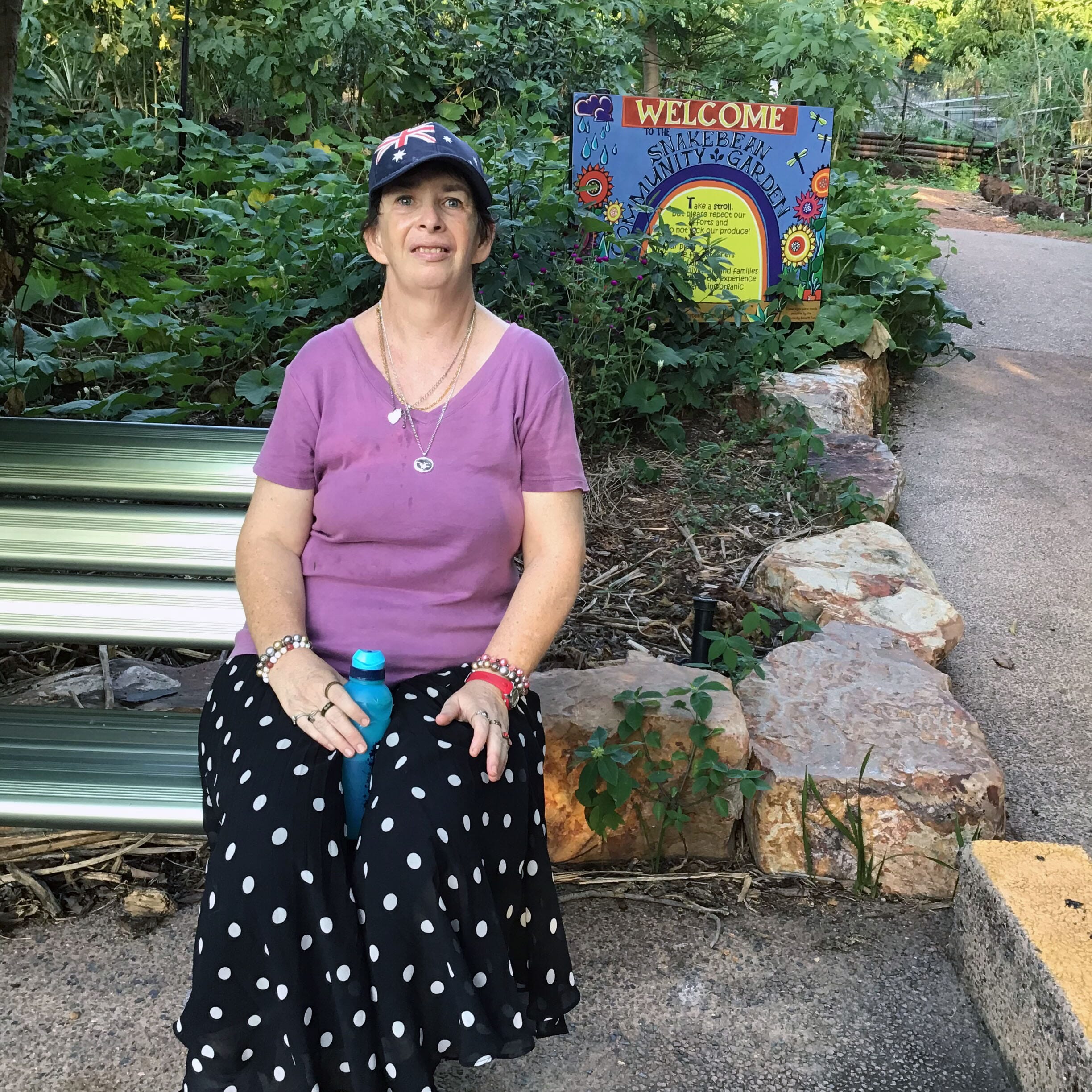 Margaret seated beside a welcome sign at a community garden in Darwin during community access.