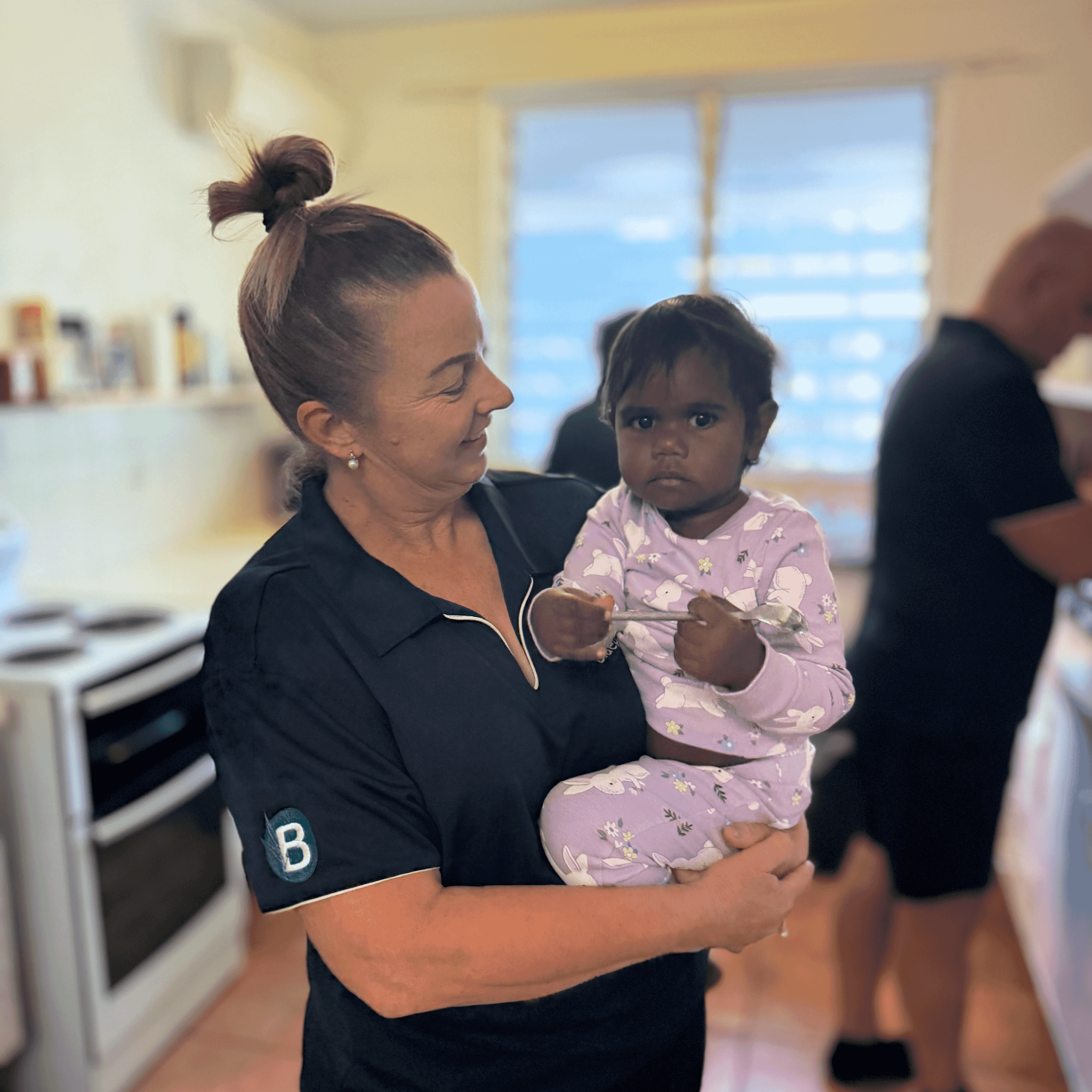 A Bluegum staff member smiles while holding a young child in a cosy kitchen, with another team member preparing food in the background.