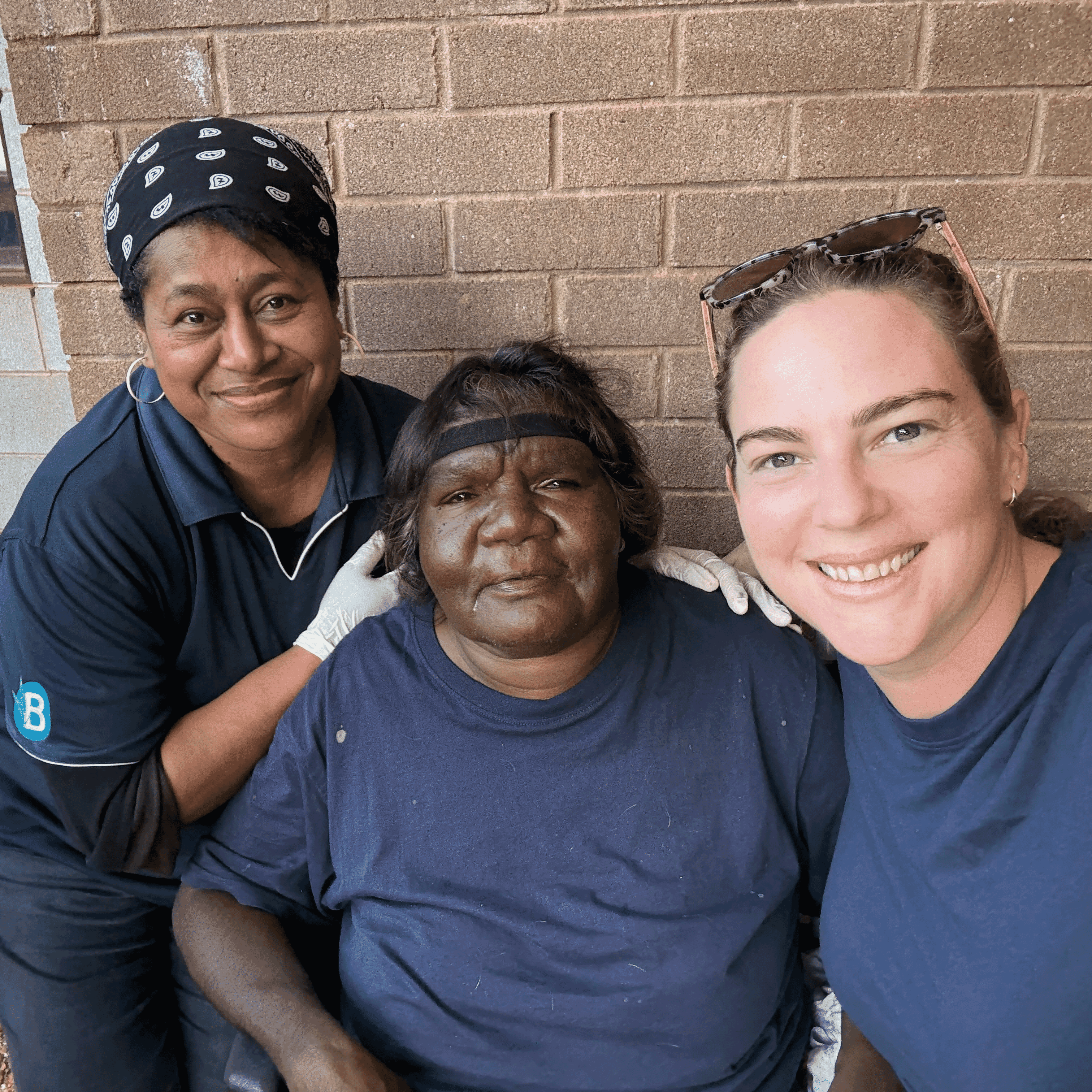 Cynthia sits between Rose and Liti, all smiling for a photo against a brick wall at the Tennant Creek Supported Independent Living house.