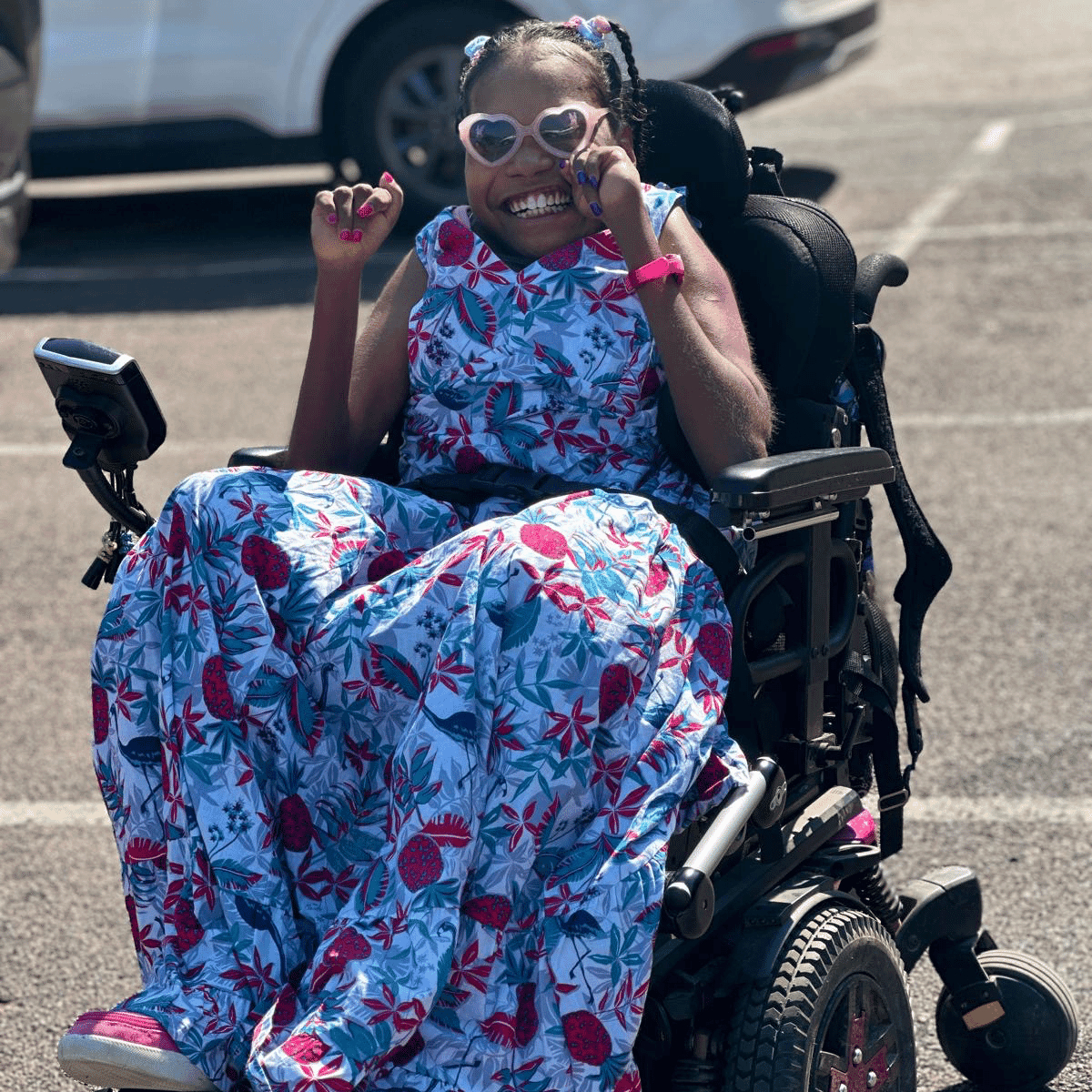 Stella smiles in heart-shaped sunglasses while sitting in her powered wheelchair, enjoying a sunny day out in Darwin.
