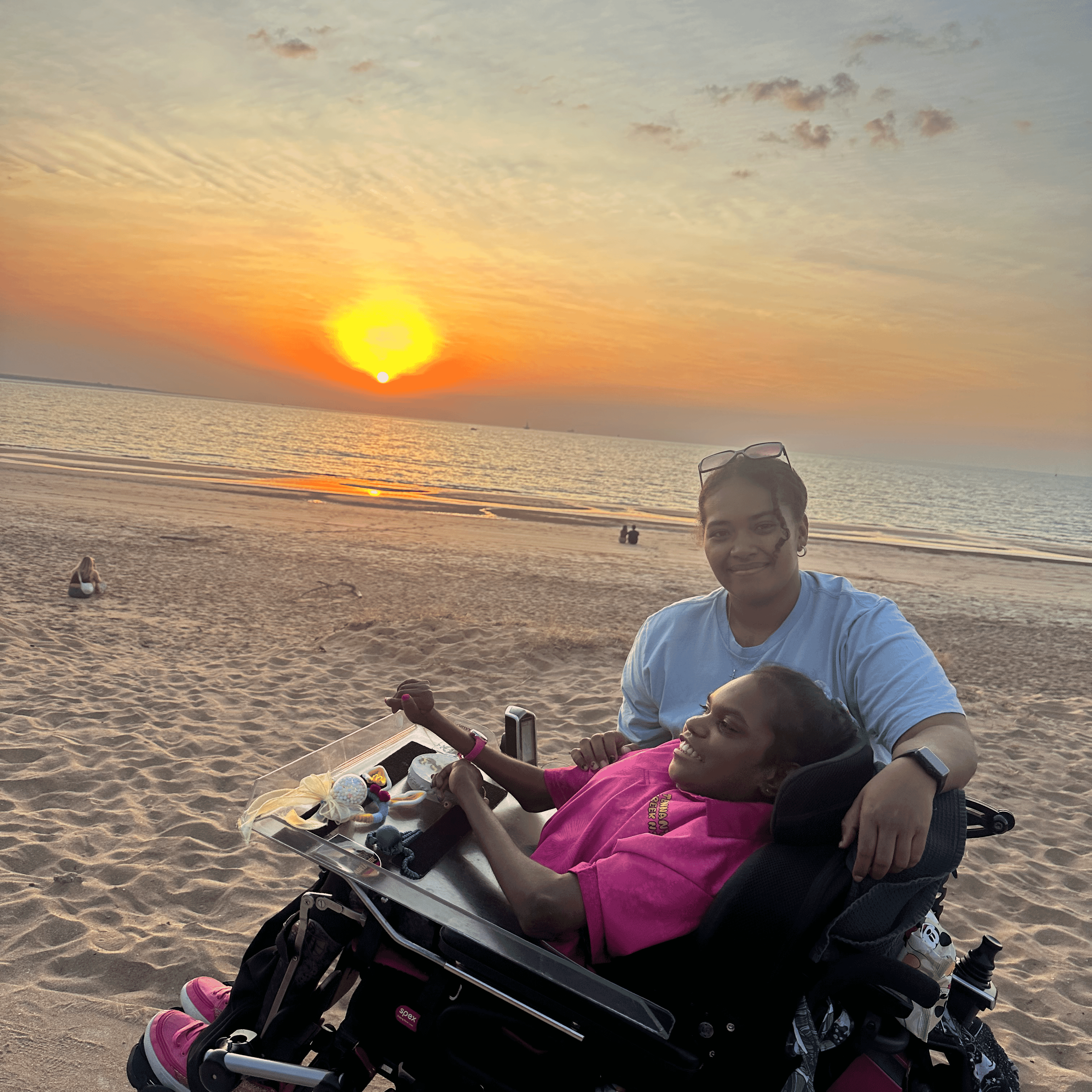 Stella smiles in her powered wheelchair while La stands beside her on the sand, watching a vivid orange sunset over the sea.