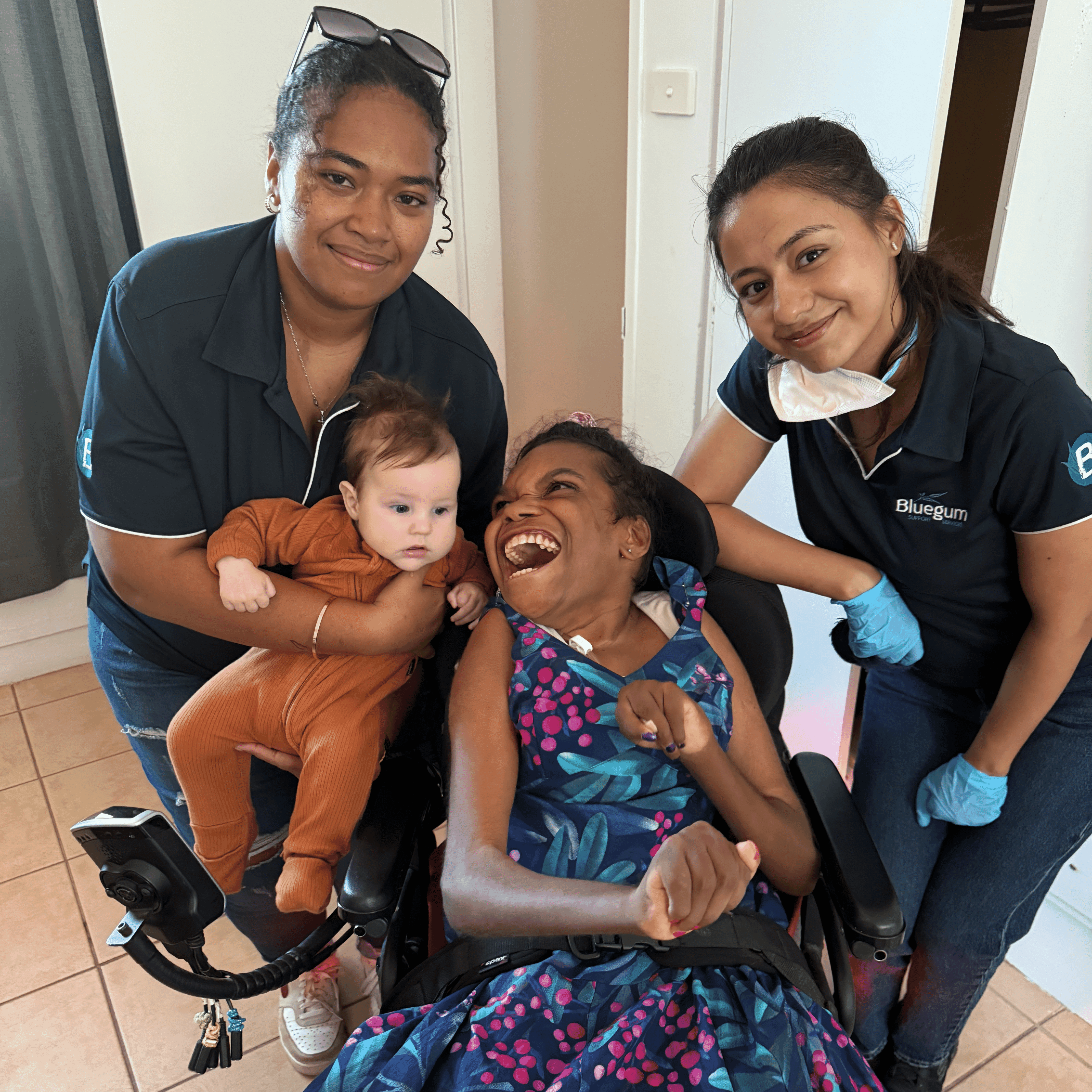 Stella smiles in her wheelchair as two Bluegum staff stand beside her—one cradling a baby—during a friendly visit in Darwin.